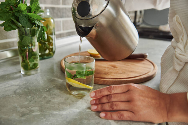 Ausgeschnittene Ansicht einer Frau im Gewand, die Wasser aus einer Teekanne in ein Glas mit Minzblättern und Zitronenscheibe gießt und gesundes Wasser mit Minze und Zitrone zubereitet. Nahaufnahme. - Foto, Bild
