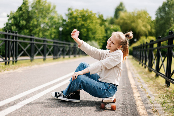 Roztomilé blondýny dospívající dívka sedí na skateboardu v městském parku chatování na smartphone s přáteli a přičemž selfie. - Fotografie, Obrázek
