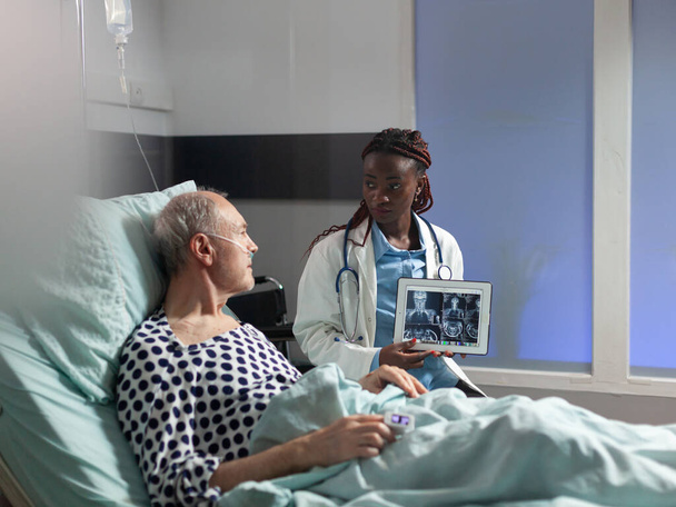 Αφροαμερικανός γιατρός κάθεται δίπλα σε ανώτερο άνδρα εξηγώντας τραύμα στο σώμα - Φωτογραφία, εικόνα