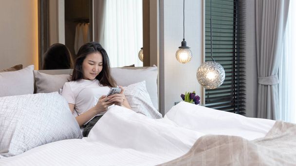 Asian woman uses mobile phone in beautiful modern bedroom at home after she wakes up in the morning, Entertaining or relaxing using mobile phone technology, Wake-up activities, Happy lifestyle. - Fotó, kép