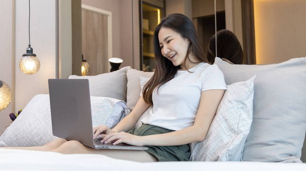 アジアの女性は、彼女が午前中に目が覚めた後、自宅で近代的な寝室でベッドの上にラップトップを使用しています,ラップトップ技術を使用して娯楽やリラックス,ウェイクアップ活動,幸せなライフスタイル. - 写真・画像