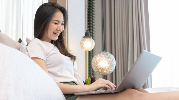 Asian woman uses laptop on bed in modern bedroom at home after she wakes up in the morning, Entertaining or relaxing using laptop technology, Wake-up activities, Happy lifestyle. - Foto, Imagem