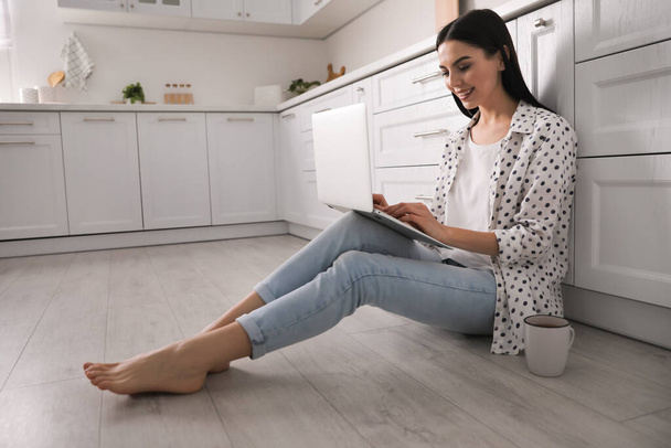 Happy woman with laptop and cup of drink sitting on warm floor in kitchen. Heating system - Zdjęcie, obraz