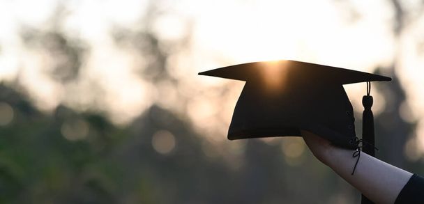 рука тримає бакалаврський капелюх з боке на фоні заходу сонця
 - Фото, зображення