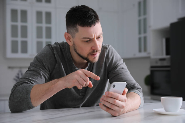 Emotional man with smartphone at table in kitchen. Online hate concept - Photo, image