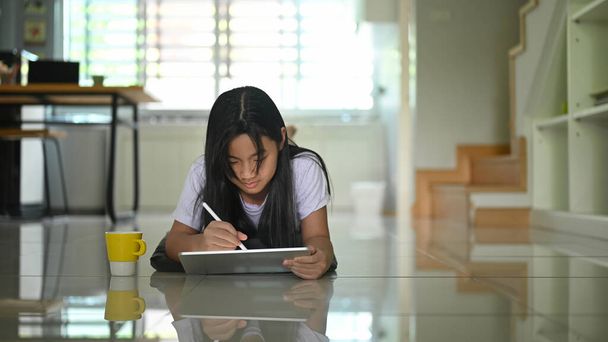 Mała dziewczynka używa tabletu komputerowego i długopisu leżąc na parterze salonu.. - Zdjęcie, obraz