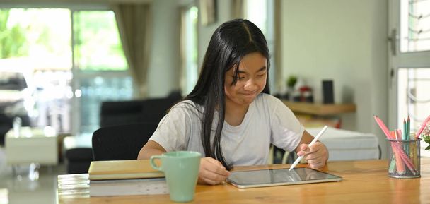 Μια μαθήτρια χρησιμοποιεί μια λευκή κενή οθόνη tablet υπολογιστή και στυλό γραφίδα στο καθιστικό. - Φωτογραφία, εικόνα