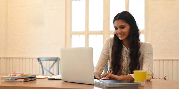 Yliopiston opiskelija opiskelee verkossa istuessaan puisen työpöydän ääressä. - Valokuva, kuva