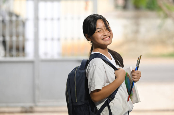 Een schoolmeisje houdt schilderspullen vast en draagt een schooltas terwijl ze staat te wachten op een schoolbus.. - Foto, afbeelding