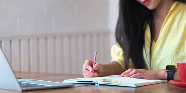 Молода жінка робить нотатки, сидячи перед комп'ютерним ноутбуком на дерев'яному робочому столі
. - Фото, зображення