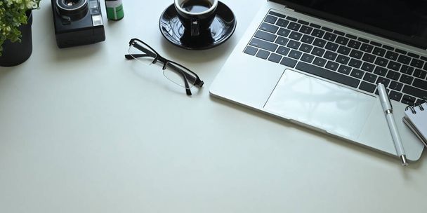 λευκό γραφείο εργασίας με φορητό υπολογιστή, στυλό, γυαλιά, φλιτζάνι καφέ, σημείωση, ρετρό κάμερα, φιλμ και γλάστρα. - Φωτογραφία, εικόνα