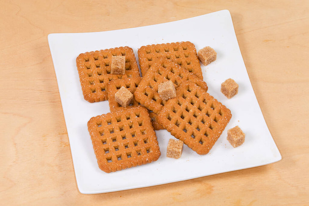 Platte knapperige vierkant gevormde koekjes, bestrooid met kristalsuiker voor het bakken en bruine suikerklontjes op een witte vierkante schaal op een houten ondergrond - Foto, afbeelding