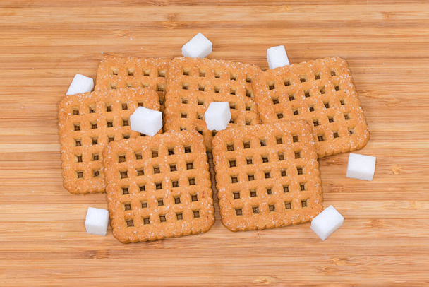 Επίπεδα τραγανά μπισκότα σε σχήμα τετραγώνου, πασπαλισμένα με ζάχαρη πριν το ψήσιμο και κύβους ζάχαρης σε ξύλινη επιφάνεια μπαμπού - Φωτογραφία, εικόνα