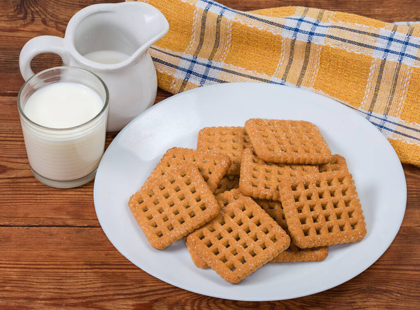 Ploché křupavé hranaté sladké sušenky na bílém talíři a mléčné výrobky na starém venkovském stole s ubrouskem - Fotografie, Obrázek
