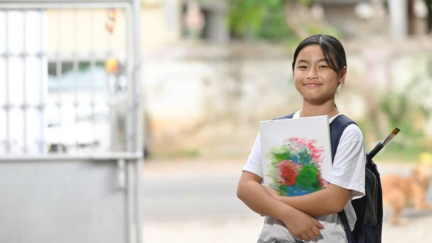 Una studentessa tiene in mano l'attrezzatura per dipingere e porta una borsa della scuola in piedi e in attesa di uno scuolabus. - Foto, immagini