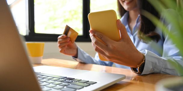 Uma mulher bonita está pagando contas via telefone celular e cartão de crédito na mesa de trabalho de madeira. - Foto, Imagem