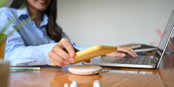 Ofisteki bir kadının kesilmiş görüntüsü bilgisayar laptopunda yazarken akıllı telefonu kablosuz şarj cihazıyla şarj ediyor.. - Fotoğraf, Görsel