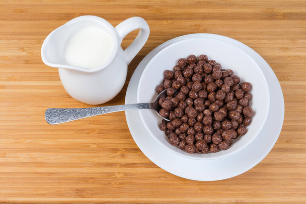Breakfast cereal chocolate balls and spoon in bowl, milk in the small milk jug on a wooden surface, top view - Photo, Image