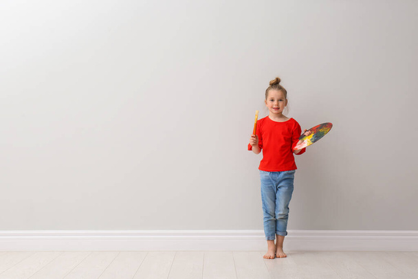 Little girl with brush and paint palette near light grey wall indoors. Space for text - Φωτογραφία, εικόνα