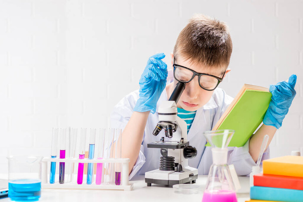 A schoolboy with a microscope and book examines chemicals in test tubes, conducts experiments - a portrait on a white background. Concept for the study of coronavirus in the laboratory - Zdjęcie, obraz