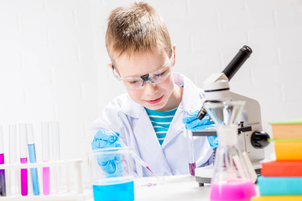 Een schooljongen met een microscoop onderzoekt chemicaliën in reageerbuizen, voert experimenten uit - een portret op een witte achtergrond. Concept voor de studie van het coronavirus in het laboratorium - Foto, afbeelding