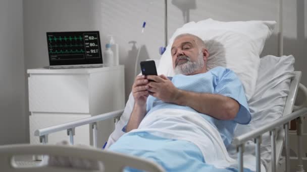 Male Patient Using Mobile Phone In The Ward At Hospital. Senior Male patient holding mobile phone while resting on bed in hospital - Filmmaterial, Video