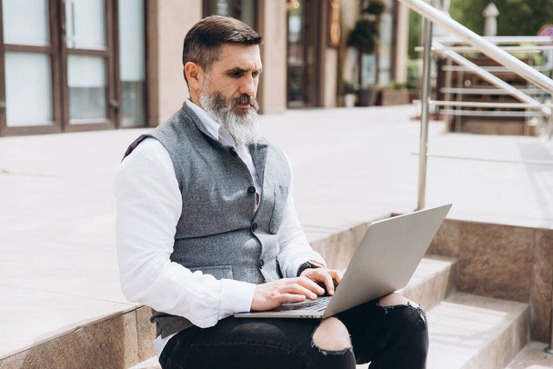 Senior mężczyzna stylowy mężczyzna z szarą brodą spędza czas na świeżym powietrzu w laptopie. - Zdjęcie, obraz