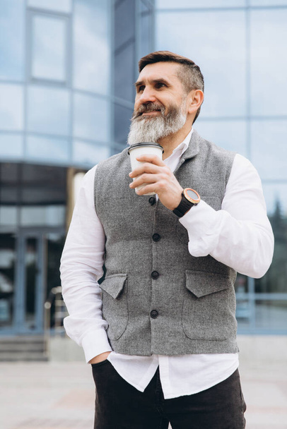 Positive brutal bearded gray-haired senior man drinking coffee on urban clean spaces - Photo, Image