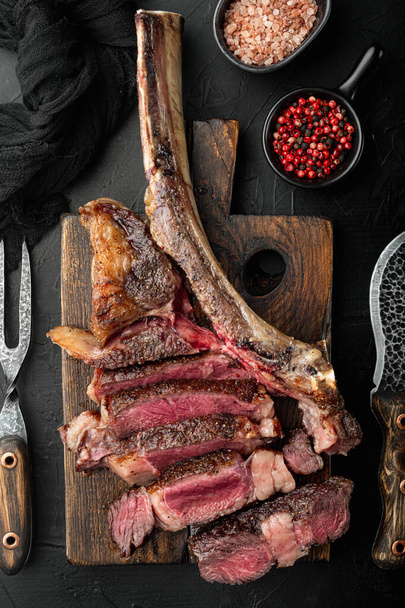 Friss grillezett hús. Grillezett marhahús steak közepes ritka készlet, tomahawk vágott, fa tálalódeszka, fekete kő háttér, felső nézet lapos fektetés - Fotó, kép