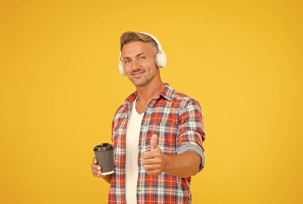 Happy man in modern earphones show thumbs up hand gesture approving and promoting takeaway coffee cup, pleasure - Zdjęcie, obraz