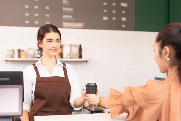 Όμορφη καυκάσια barista γυναίκα δώσει takeaway φλιτζάνι καφέ στον πελάτη και τον πελάτη που λαμβάνουν φλιτζάνι καφέ, με επίκεντρο το πρόσωπο της γυναίκας barista. Η Barista εργάζεται σε επιχειρήσεις παροχής υπηρεσιών τροφίμων και καφέ. - Φωτογραφία, εικόνα