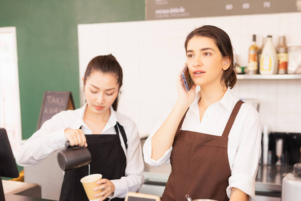 Caucasian barista woman confirms online orders with customer via mobile, Asian barista woman fills white milk into takeaway coffee cup at cafe bar. Barista work at coffee bar and food service business - Photo, Image