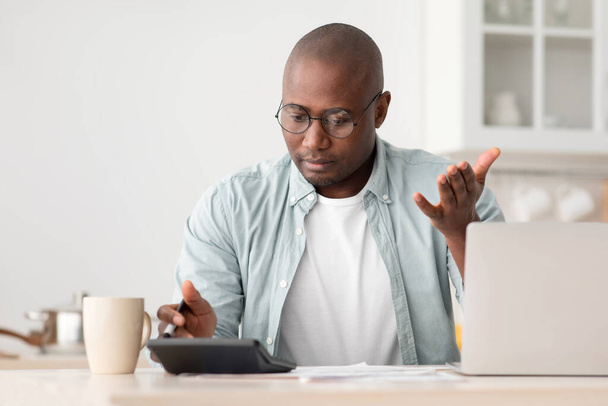 Savings and finances concept. Shocked black mature man calculating bills, using calculator, sitting in kitchen - Photo, image