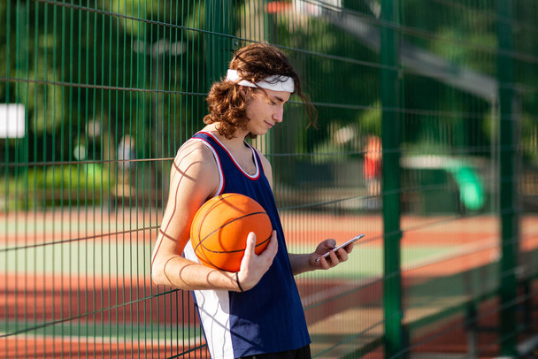 Young Caucasian basketball player using cellphone during game break at outdoor court - Zdjęcie, obraz