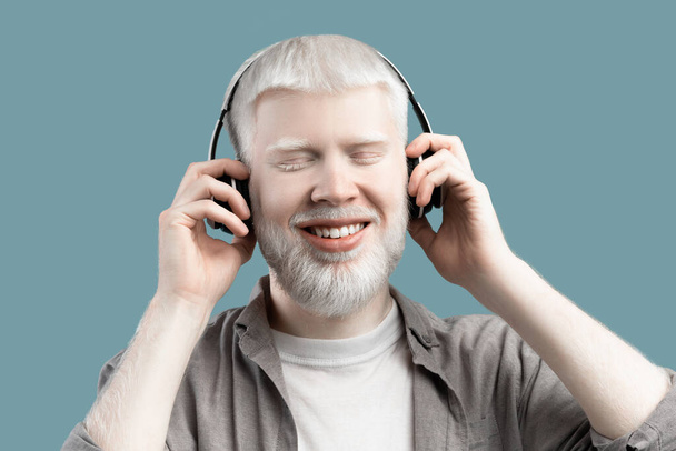 Щасливий альбіно чоловік слухає музику в бездротових навушниках і торкається їх на бірюзовому студійному фоні
 - Фото, зображення
