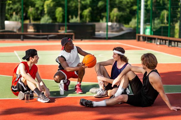 Team of young basketballers discussing their game strategy, sitting at outdoor stadium after training, free space - Fotografie, Obrázek