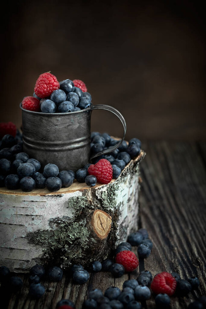 Fresh berries raspberries and blueberries in a cup. The berries are scattered on a birch stump. Birch cut. Vintage metal cup. Rustic style. Postcard. Poster. - Photo, Image