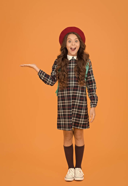 child in pupil uniform presenting product. kid in french beret on yellow background. stylish teen college student. little girl with backpack going to school. education concept. school and fashion - Fotoğraf, Görsel