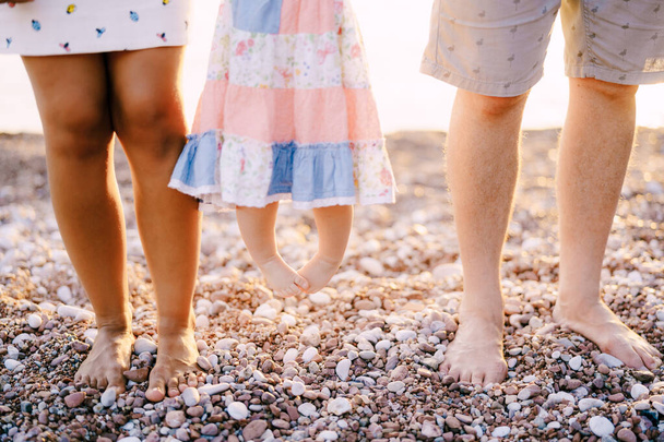 Legs of mom, dad and little daughter in a dress standing on a pebble beach  - Foto, Imagen