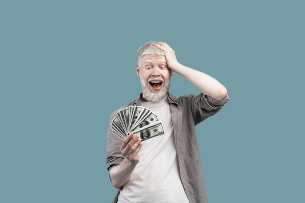 Excited extraordinary albino guy holding lots of money, touching head with shocked emotion over turquoise background - 写真・画像