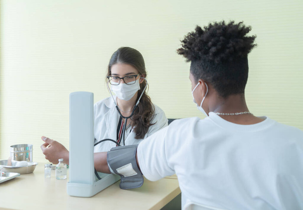 Женщина-врач в маске для лица, проверяет кровяное давление больного молодого чернокожего афроамериканца, больного в клинике или больнице в области здравоохранения и медицинского лечения. Людей. Коронавирус. - Фото, изображение