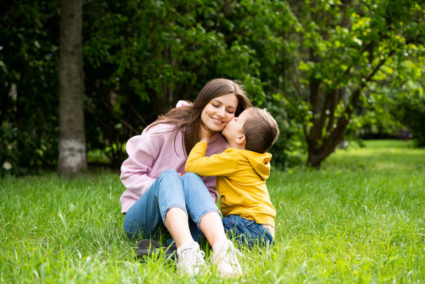 Young woman mom sits on green grass in summer park with boy son. Son kisses mom's cheek. - Photo, image