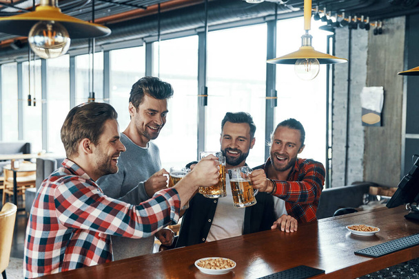 Happy young men in casual clothing toasting each other with beer and laughing while sitting in the pub - Photo, image