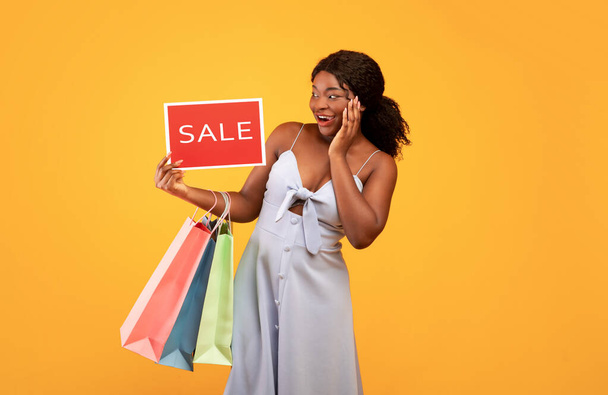 Pretty African American woman holding shopping bags and SALE sign, feeling excited over orange studio background - Foto, immagini