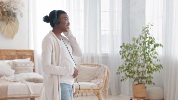 Šťastná africká americká nastávající matka těhotná afro černošky etnická žena tanec doma poslech hudby se sluchátky těší píseň melodie vzlétá staví sluchátka na břicho bříška nenarozeného dítěte - Záběry, video
