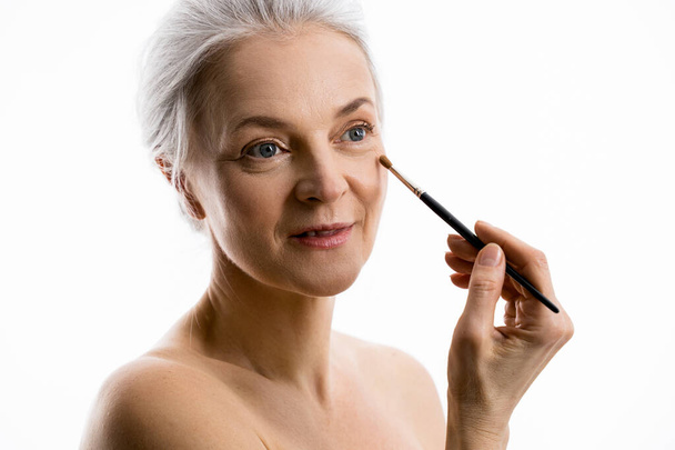 Mature woman is using brush while taking care of her face isolated on white background - Zdjęcie, obraz