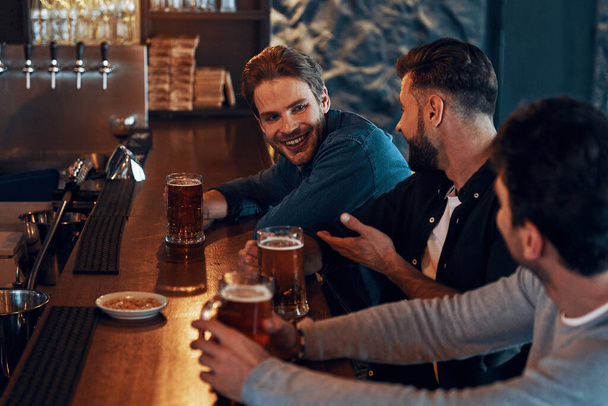 Happy young men in casual clothing drinking beer and bonding together while sitting in the pub - Foto, Imagem