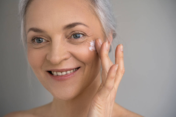 Woman putting cream on facial skin and smiling - Photo, image