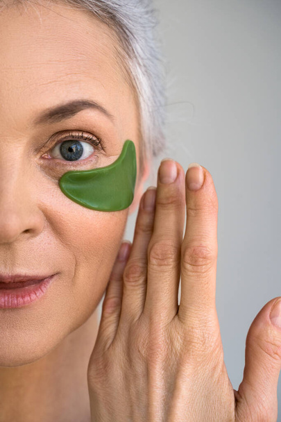 Mature woman applying cosmetic eye patches while standing at the studio - Foto, Imagem