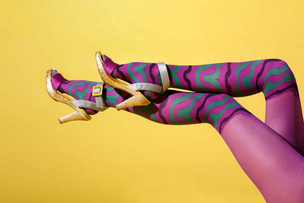 Sexy piernas bien formadas de mujer en multicolor colante púrpura, media corporal o pantimedias con zapatos de plataforma de aguja elegantemente cruzados sobre un fondo de estudio amarillo con copyspace - Foto, Imagen